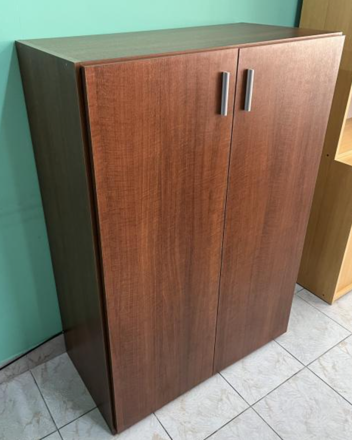 Armario de madera de dos puertas con tiradores metálicos sobre un suelo de baldosas y una pared verde.