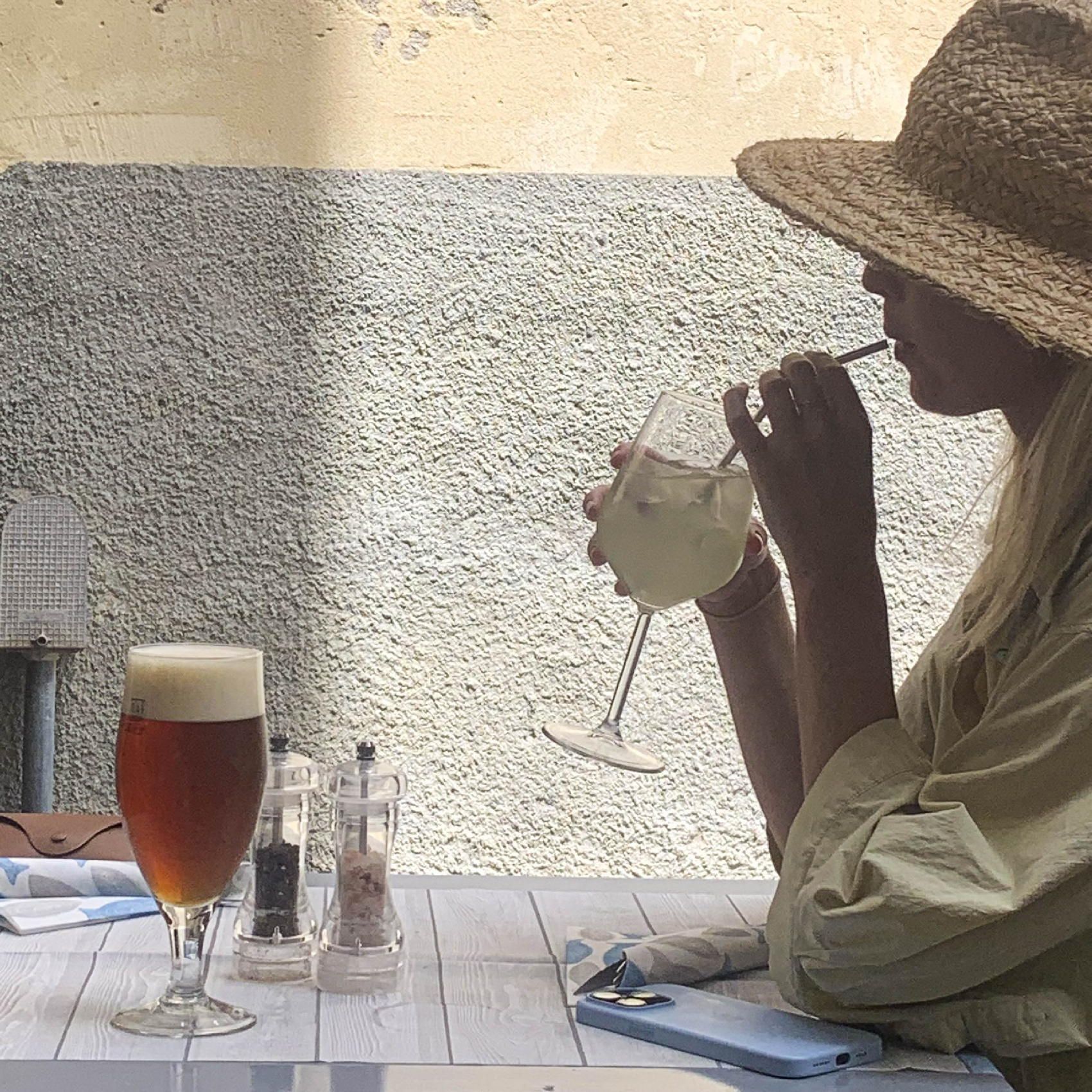 Una persona con un cappello di paglia sorseggia una bevanda a un tavolino di un caffè all'aperto, accanto a una birra.