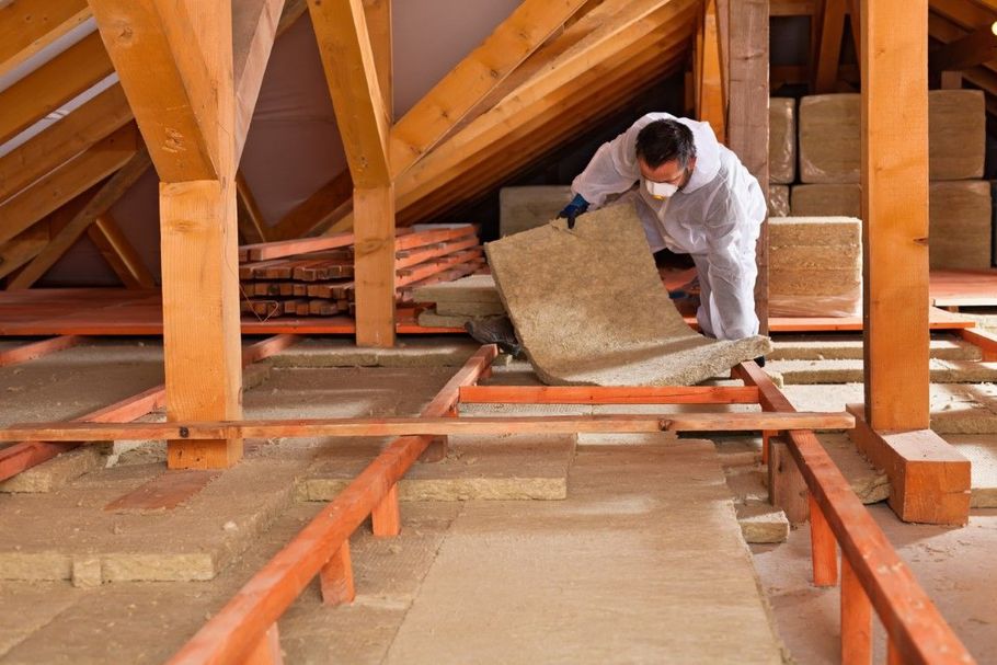 Worker installing insulation in an attic, wearing white coveralls and blue gloves.