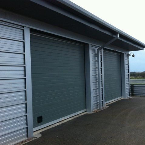 A view of a modern industrial building with two large, gray, horizontal-slat rolling garage doors.