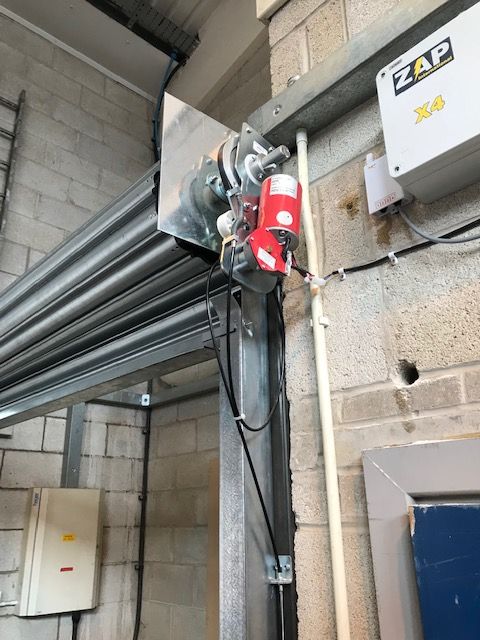 A motorized roller shutter door mechanism mounted above a door frame with a ZAP brand control box on the wall.