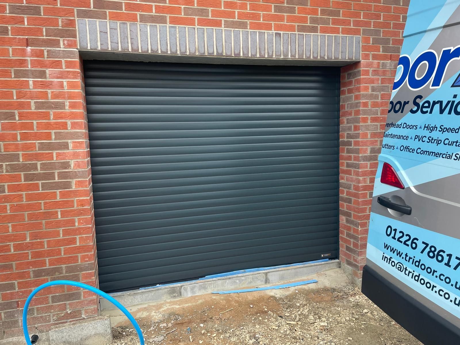 A newly installed dark gray roller garage door set in a red brick wall, next to a service van with contact information.