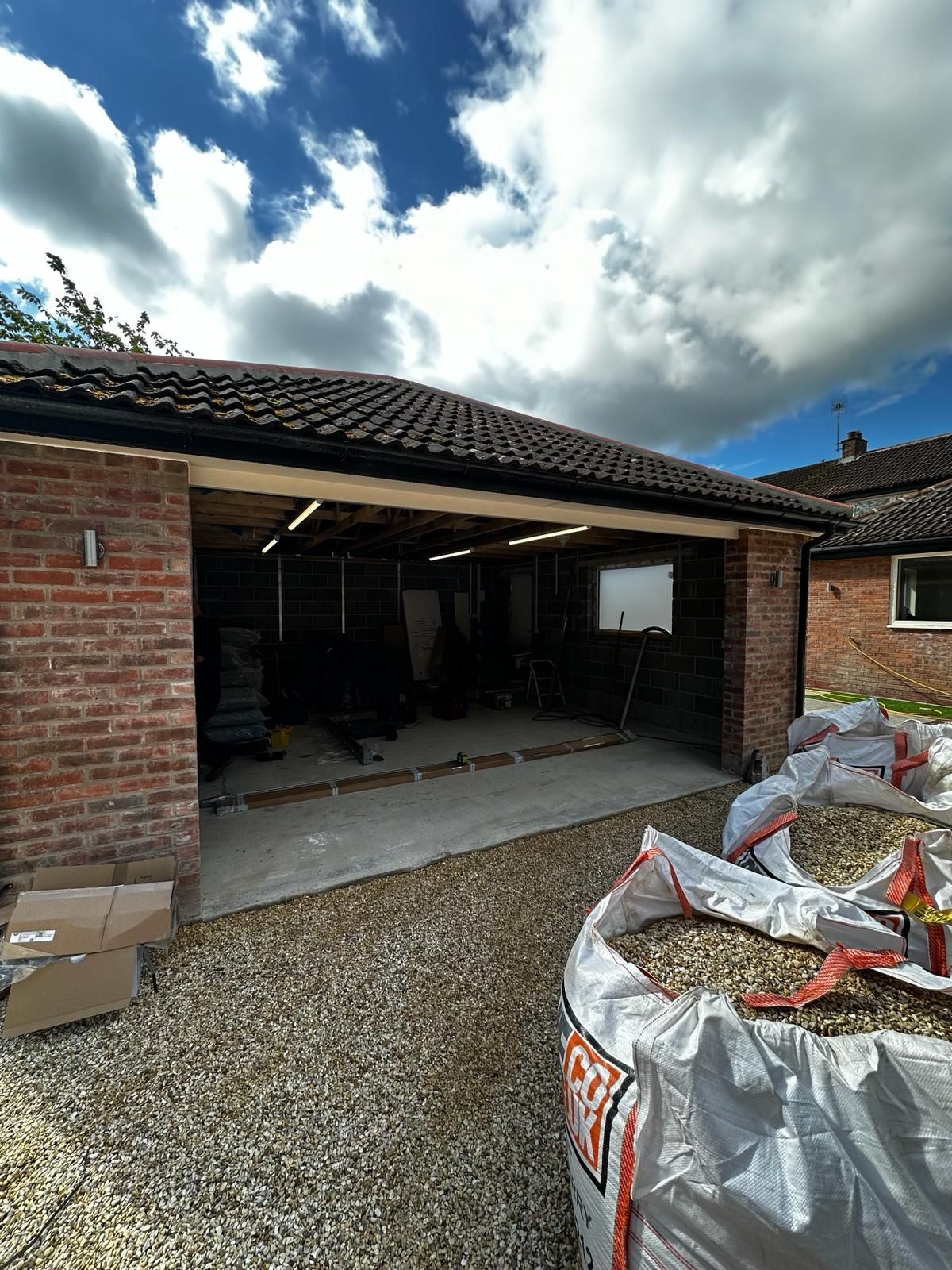 A red brick garage with an open bay under a tiled roof, with large gravel bags in the foreground on a sunny day.