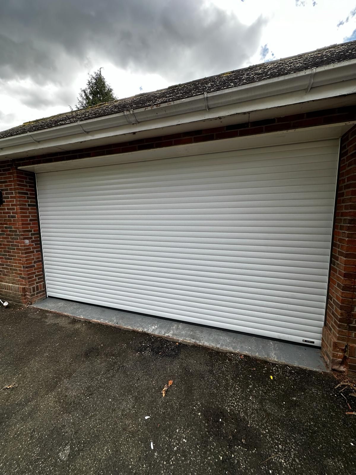 A white, slatted roller garage door installed between two brick pillars under a shingled roof.