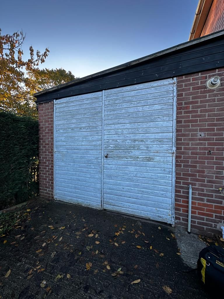 A brick garage with faded white, textured metal double doors, positioned next to a green hedge and trees.