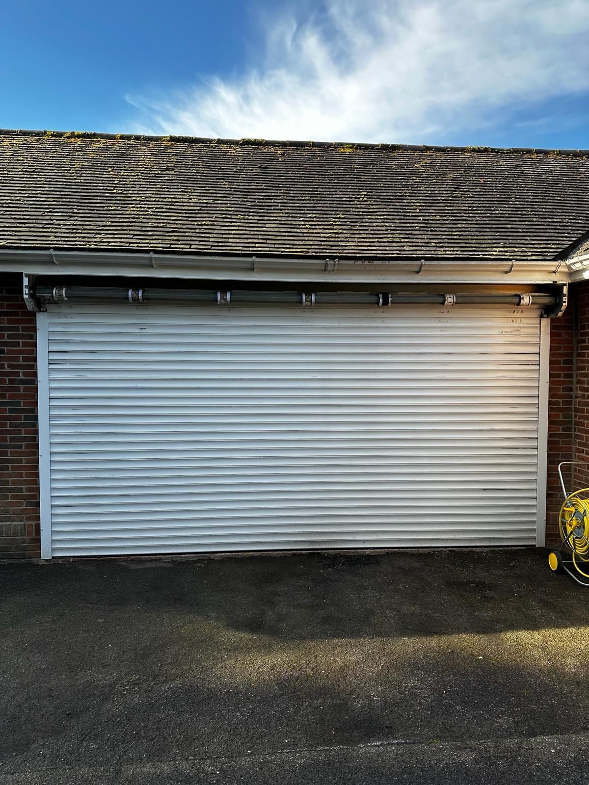 A white, horizontal-slat roll-up garage door set in a brick frame under a shingled roof.