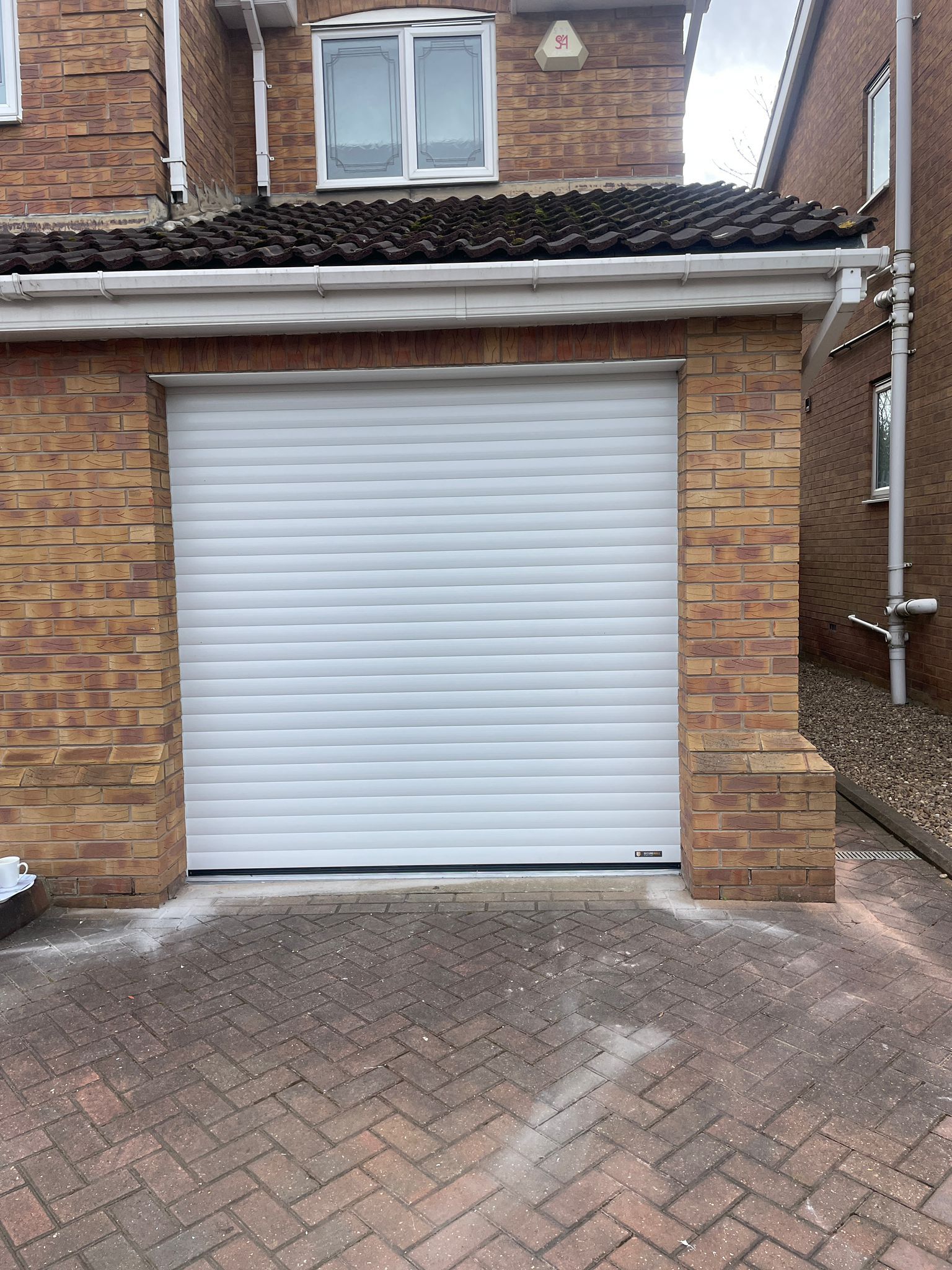 A white, horizontal-slat roller garage door set into a brick exterior wall above a paved driveway.