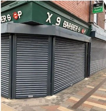 The storefront of X9 Barber Shop on a street corner, featuring dark green signage and grey closed security shutters.