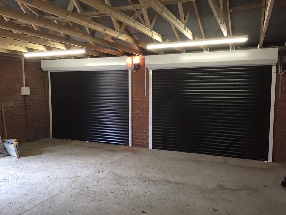 Two black, horizontal-slat roller garage doors installed side-by-side inside an unfinished brick garage.