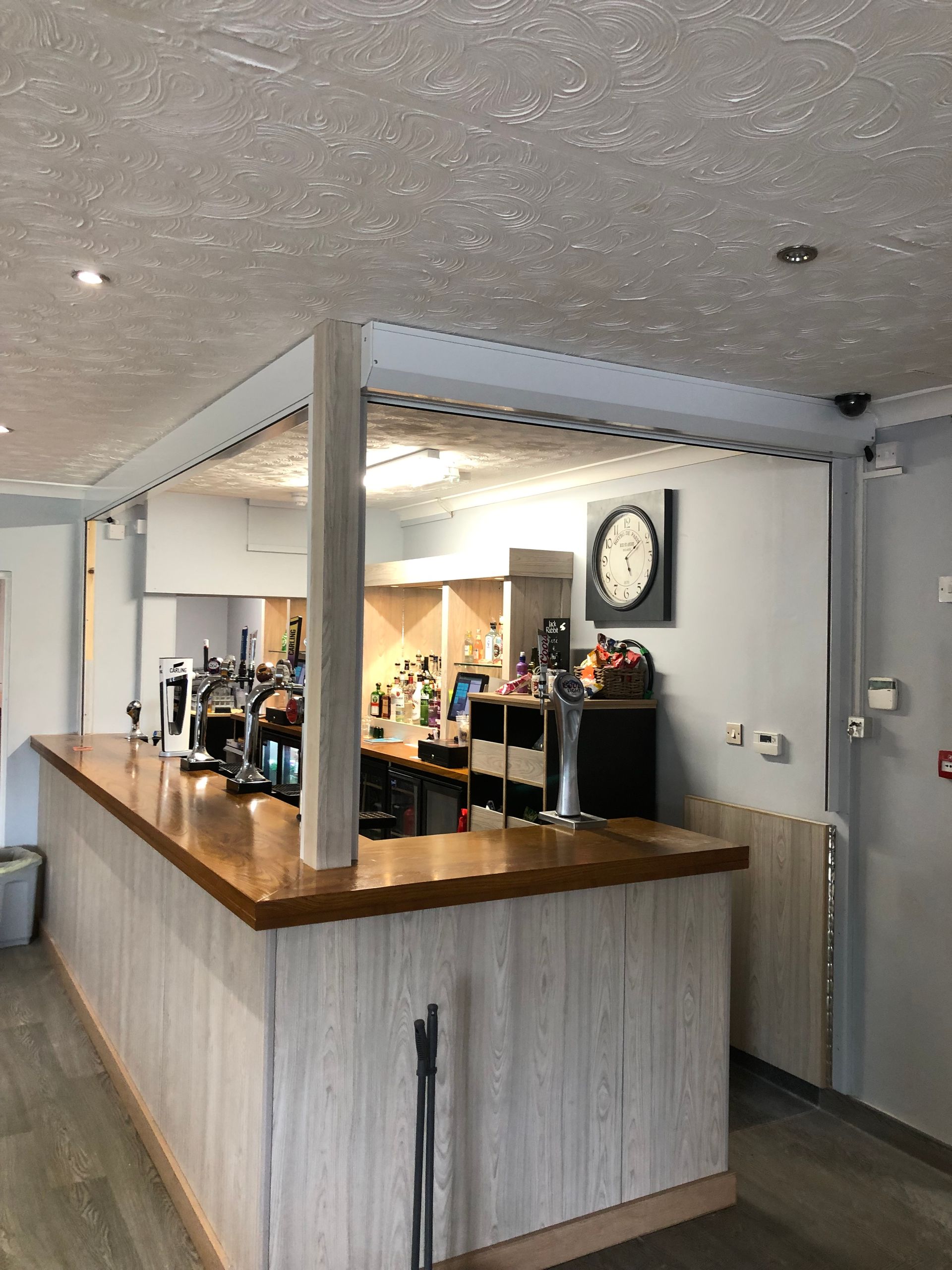A bar area with a wooden counter, draft beer taps, light-colored wood-paneled base, and a wall clock on a grey wall.