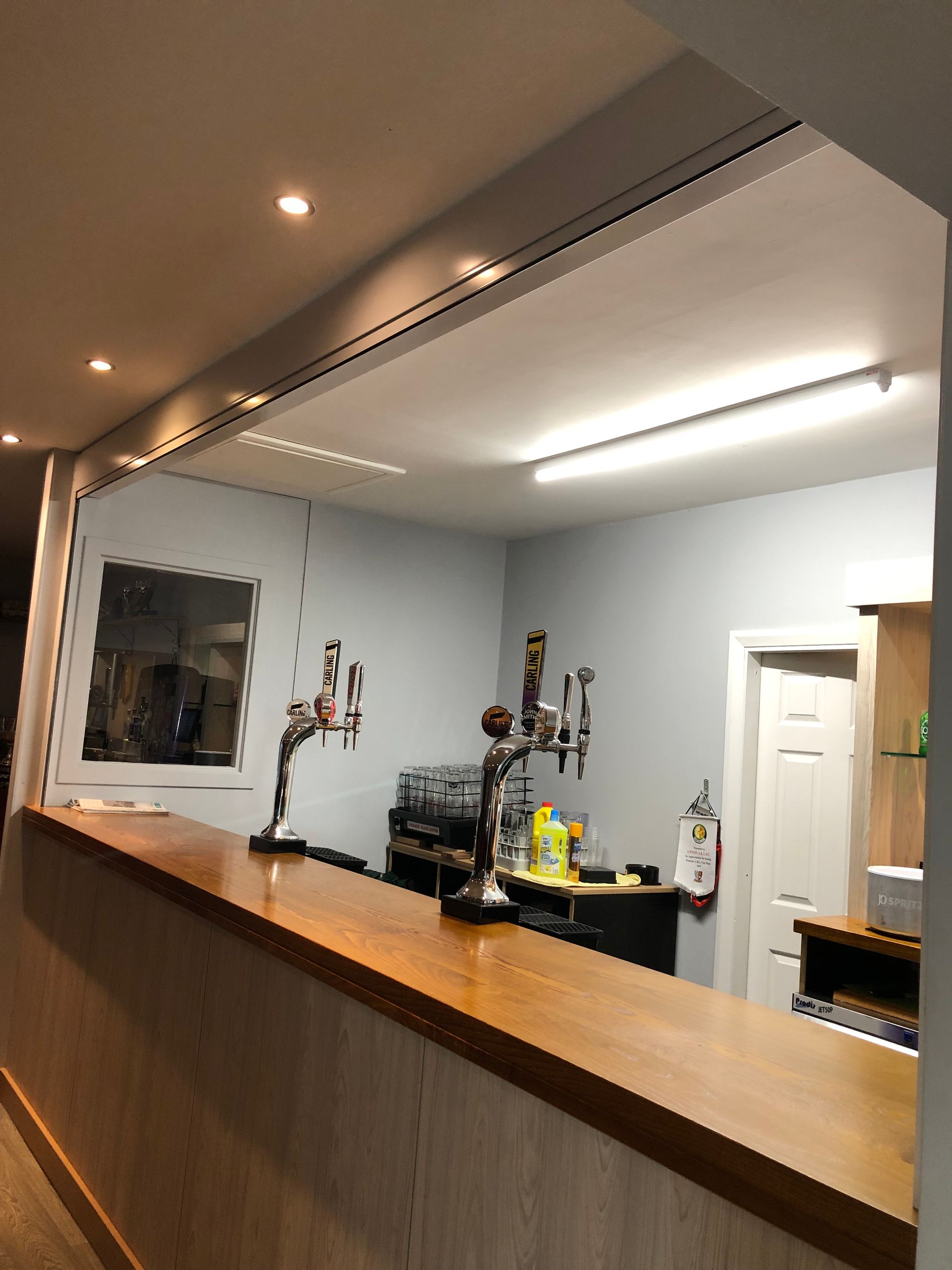 A wooden bar counter with two beer taps under bright lighting inside a room with light blue walls and a white door.