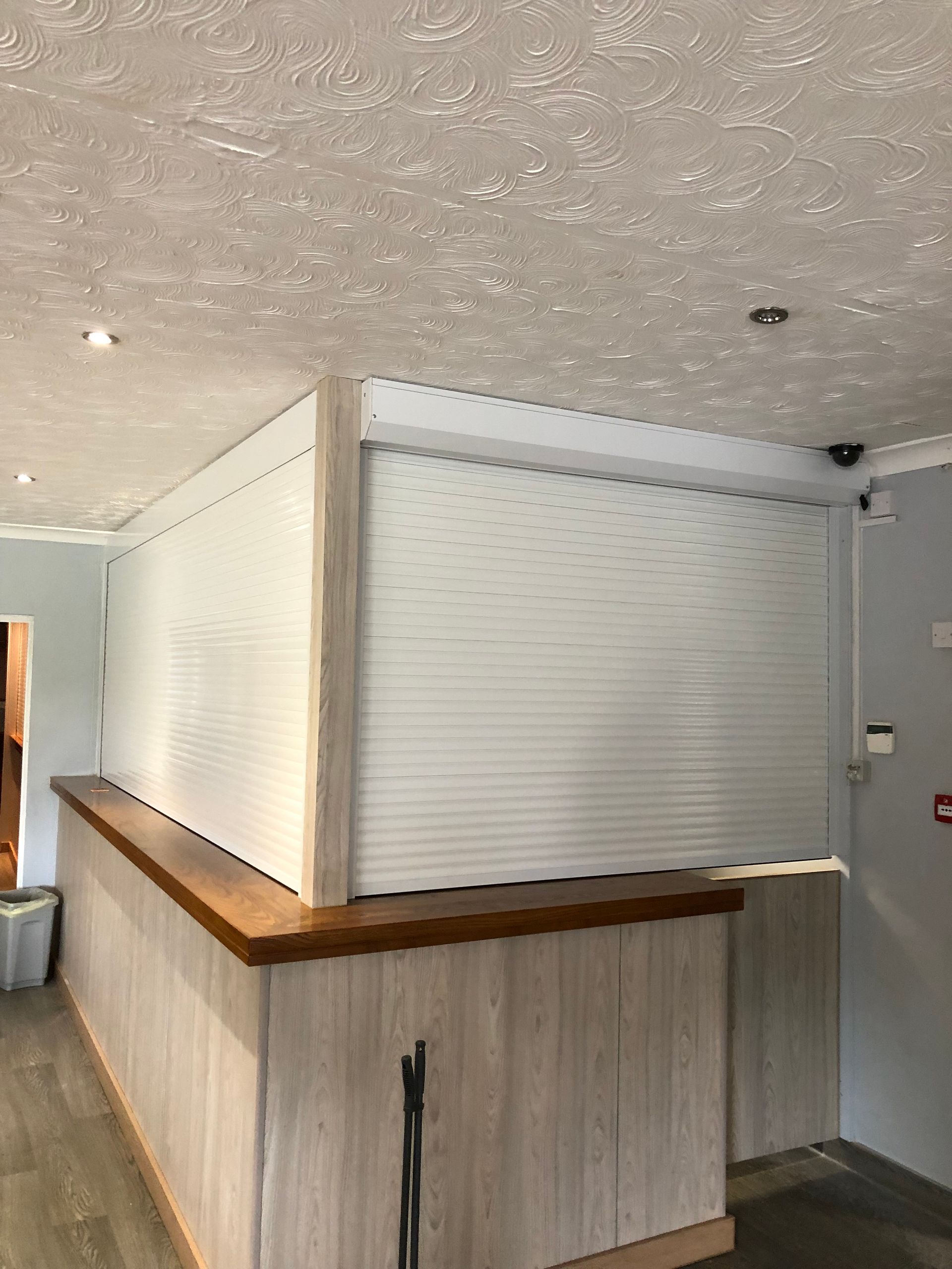 A wooden reception desk featuring a closed white roller security shutter in an indoor setting with a textured ceiling.