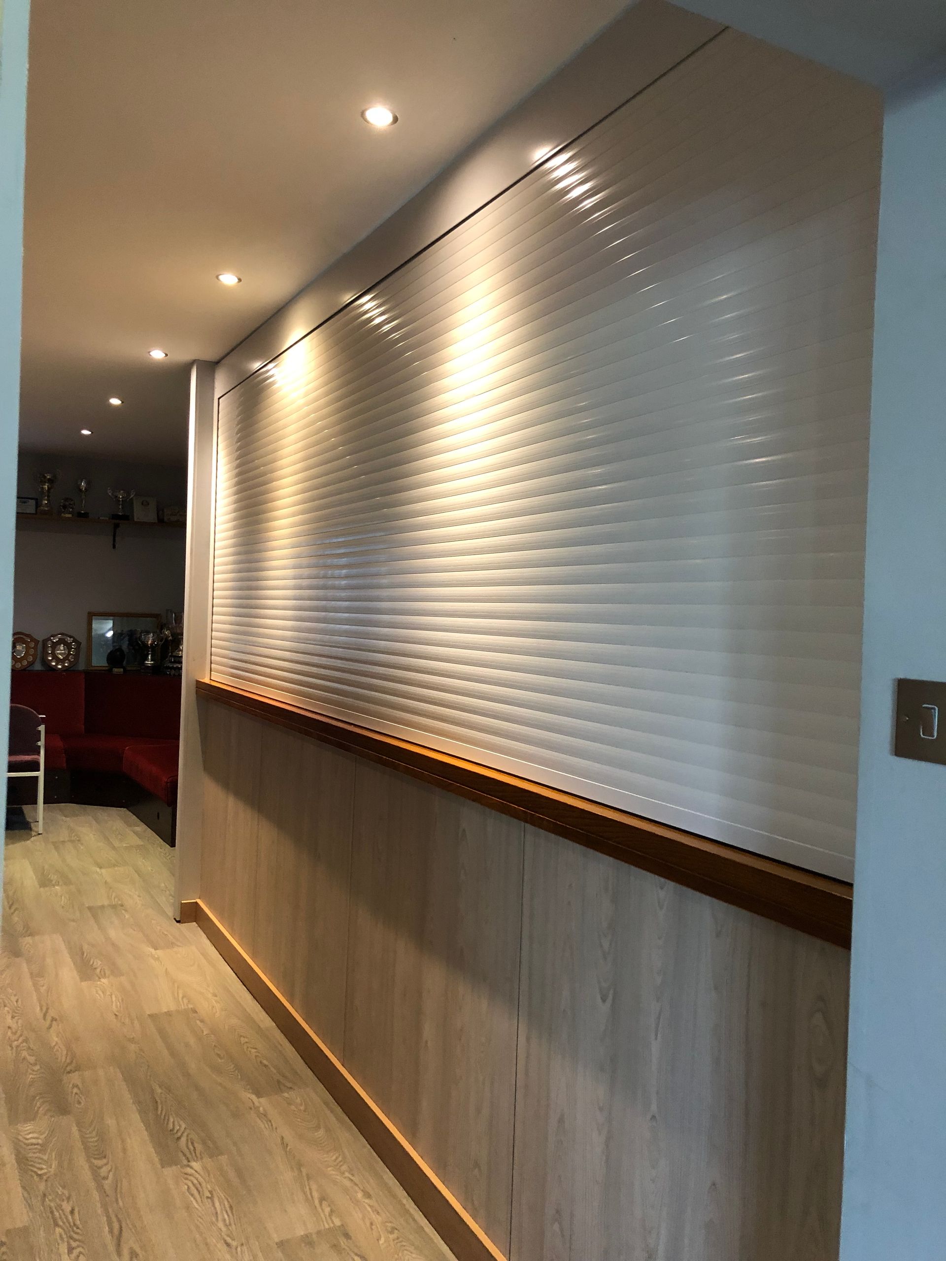 A hallway featuring a white, horizontal-slat rolling shutter covering a wall section above light wood-grain paneling.