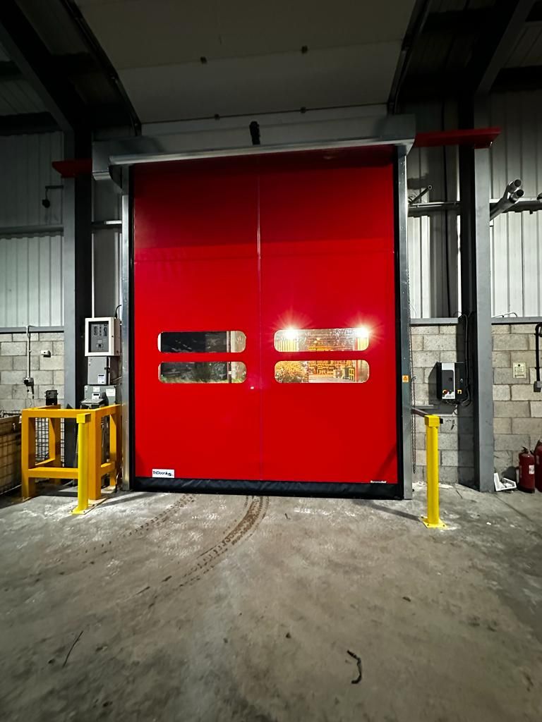 A red industrial high-speed door with horizontal vision windows installed in a warehouse with concrete flooring.