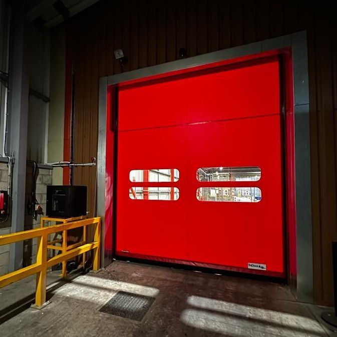 A bright red industrial rollup door with two clear vision panels, set in a metallic frame inside a warehouse facility.