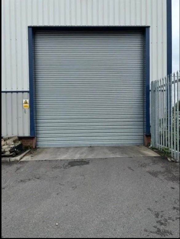 A closed grey corrugated metal roller shutter door on a light-colored industrial warehouse building.