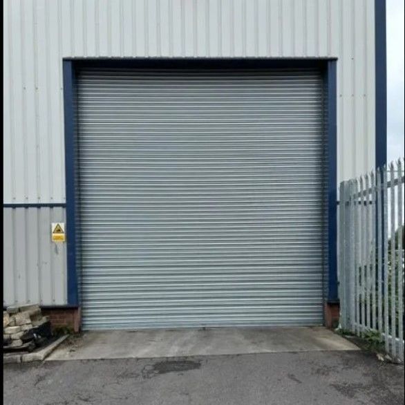 A grey industrial roller shutter door set in a white corrugated metal building wall next to a metal security fence.