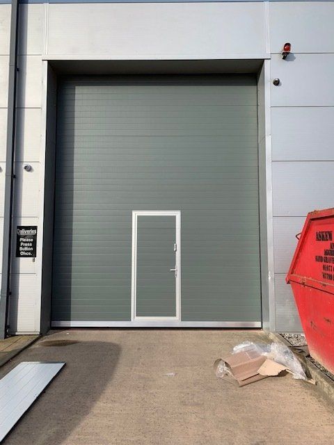 A large grey industrial roll-up door with a pedestrian pass door integrated into the center on a concrete loading bay.