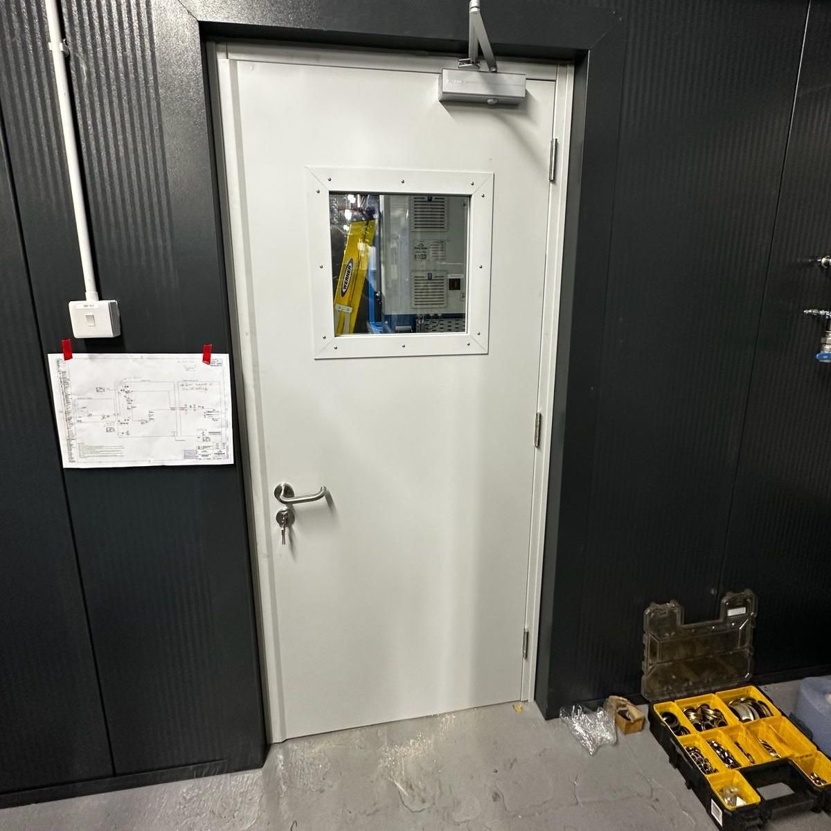 A white metal door with a small square window and a closer mechanism, centered on a dark wall with a paper note attached.