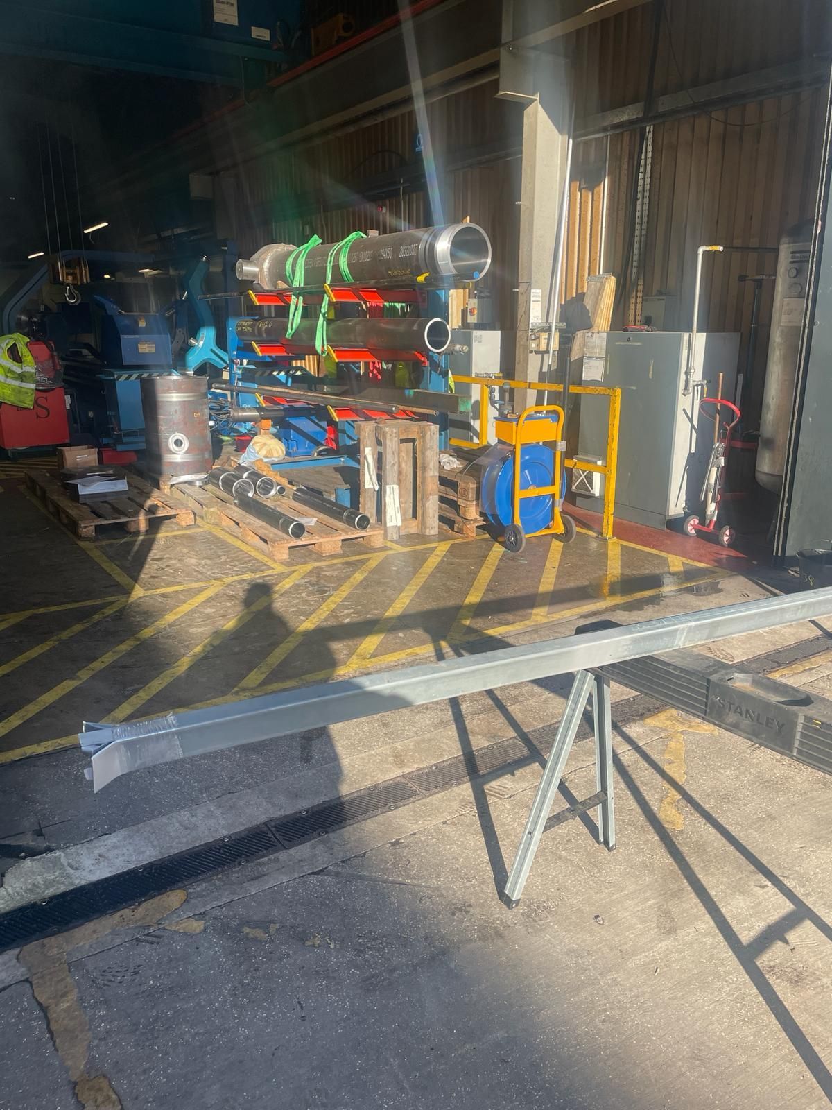 A metal beam rests on a sawhorse in the foreground of an industrial warehouse, with pipes and equipment in the background.
