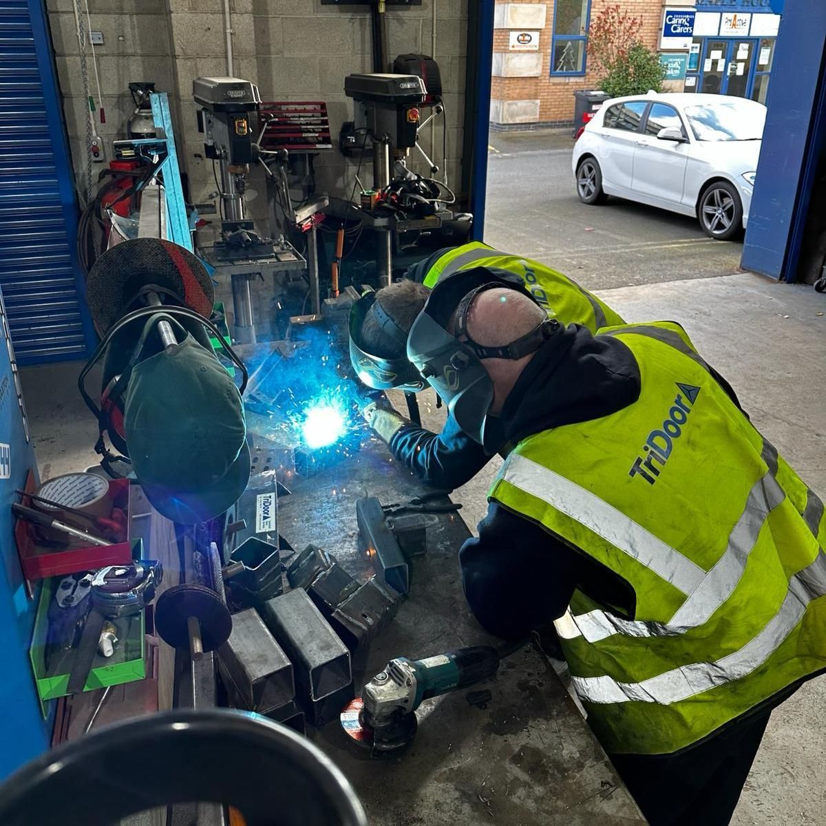 Two welders in high-visibility vests work on metal components inside a workshop, with a white car visible outside.