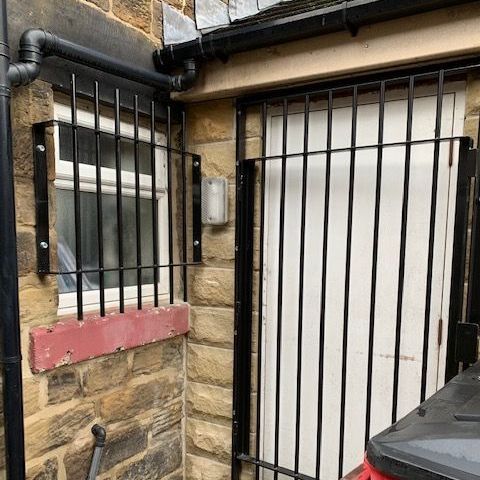A white door and a small window protected by black metal security grilles on a stone building exterior.