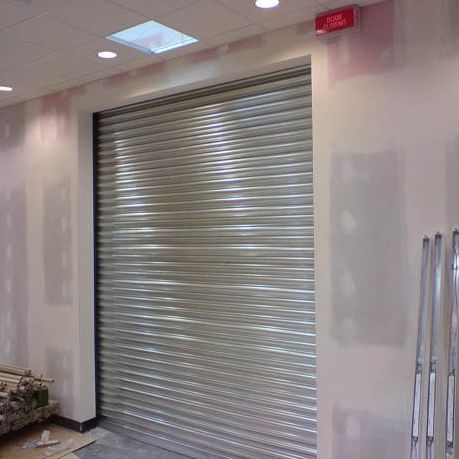 A silver metal roll-down security door installed in a room with unfinished drywall walls and a drop ceiling.