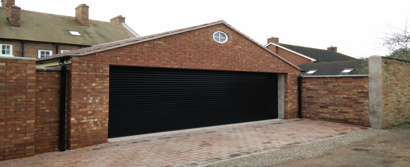 A detached brick garage with a wide black roller shutter door, situated on a paved driveway near older residential homes.