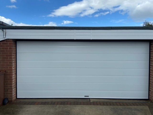 A white, horizontally paneled garage door set in a brick exterior wall under a blue sky with light clouds.