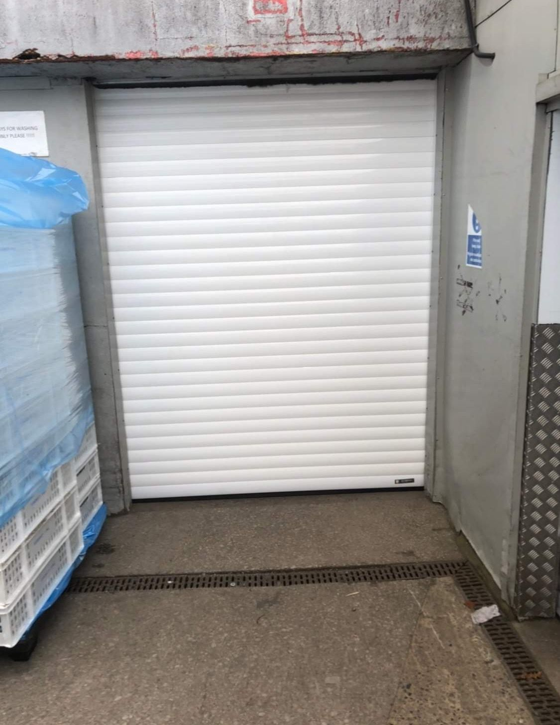 A closed white roller shutter door in a loading bay with a blue-wrapped pallet to the left.