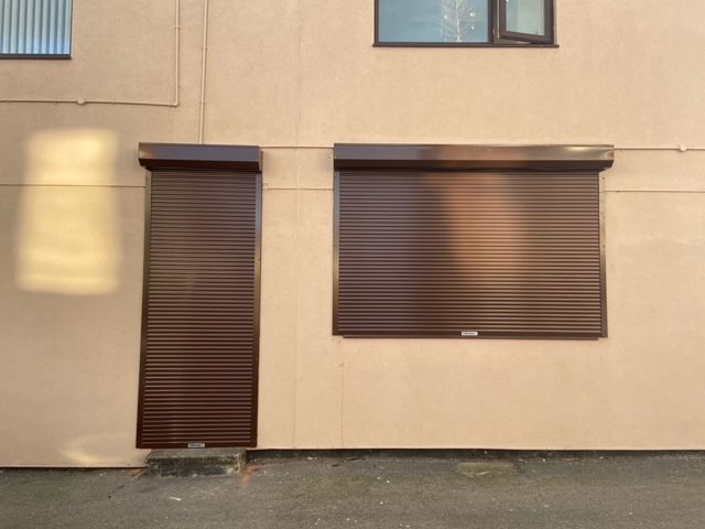 Exterior view of a beige building wall featuring a tall narrow door and a wider window covered by brown metal shutters.