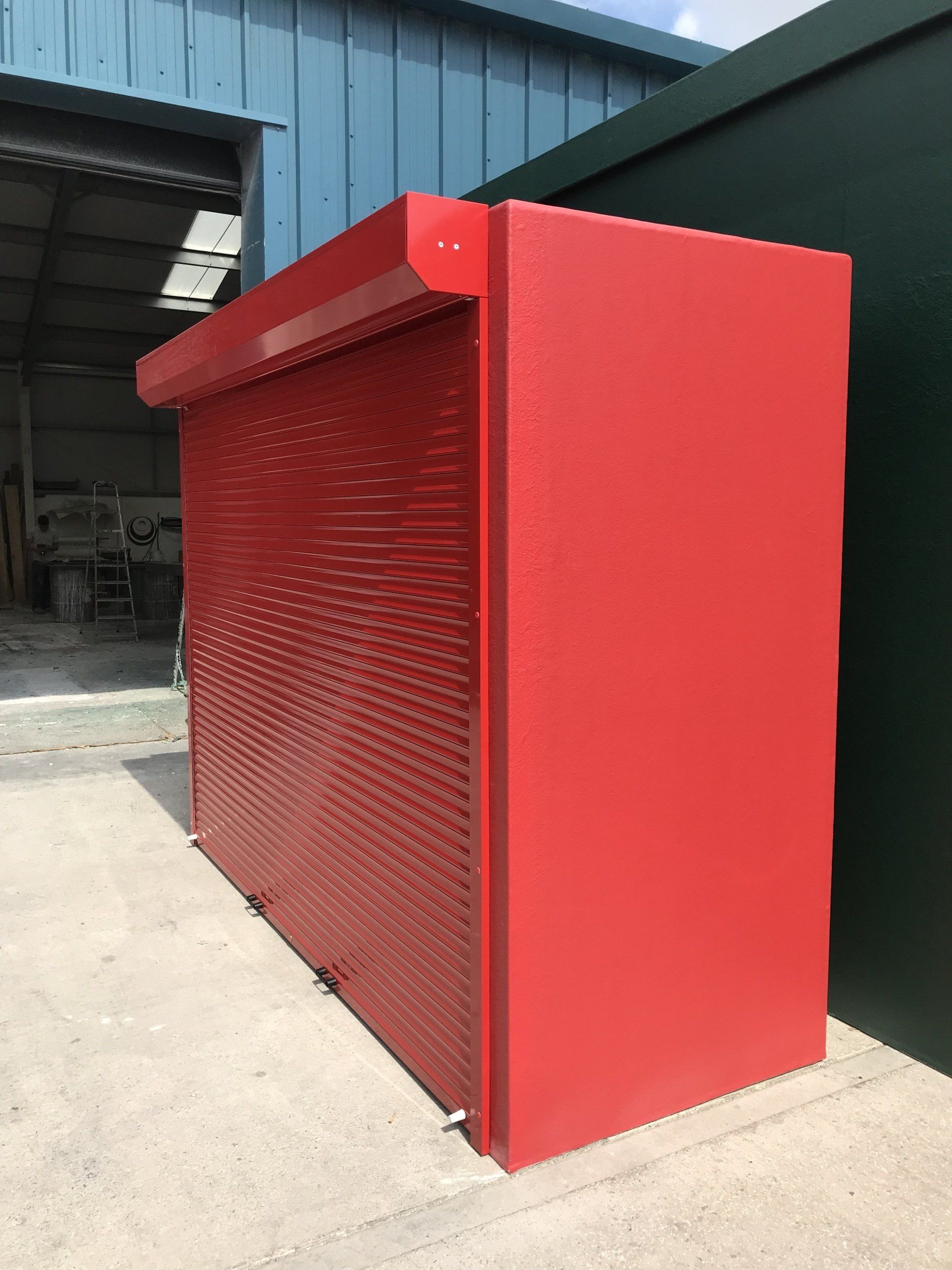 A red metal enclosure featuring a perforated, roll-down style front panel, situated outdoors on a concrete surface.