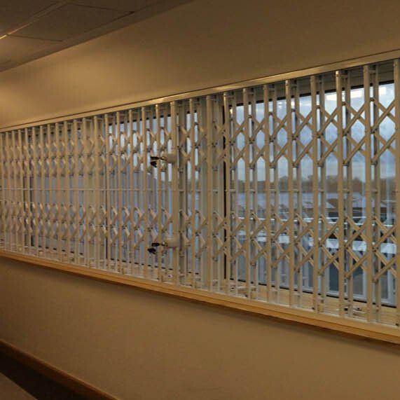 A white, retractable metal security gate installed over a long window in an indoor hallway.