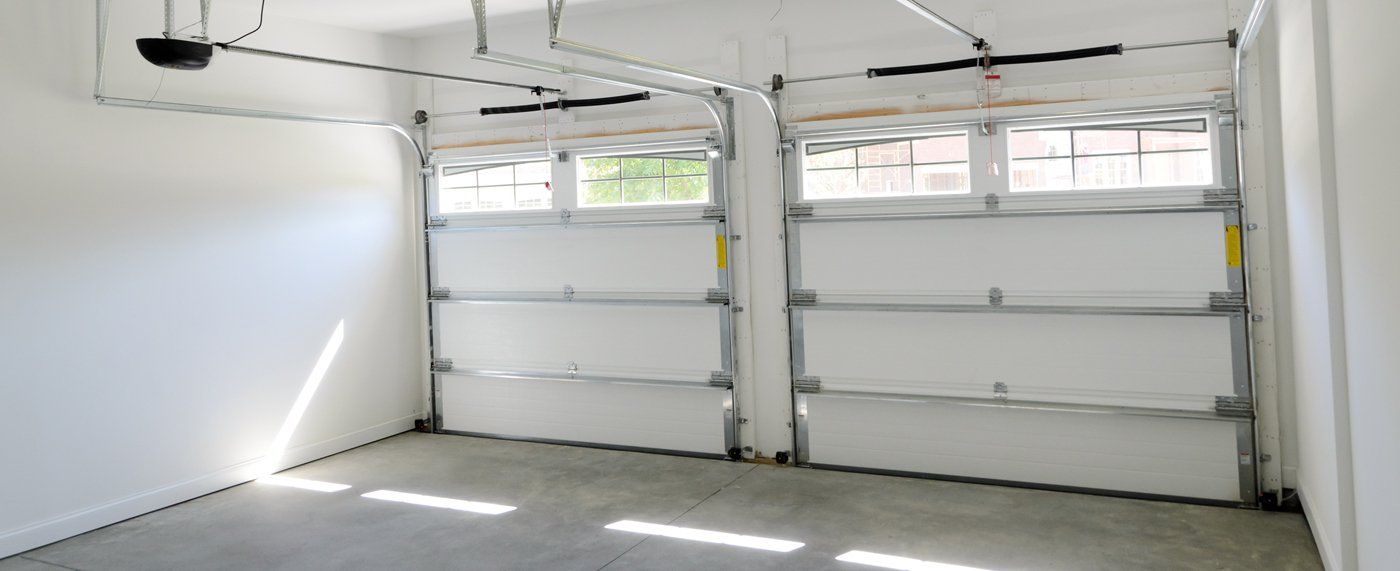 An empty residential garage with white walls, a concrete floor, and two white sectional overhead garage doors with top-row windows.