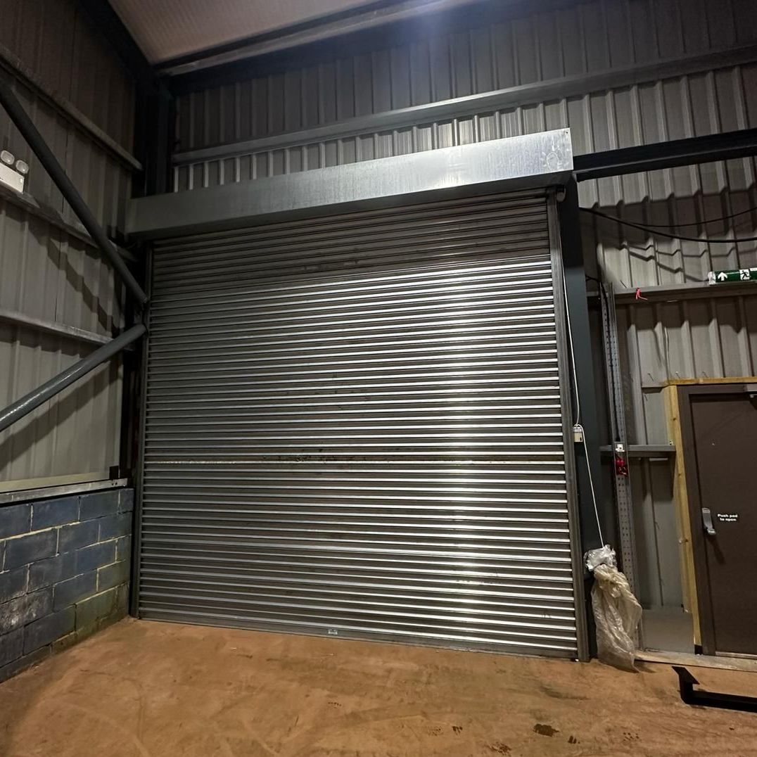 A closed metal roller shutter door inside an industrial building with grey corrugated walls and a wooden-framed side door.