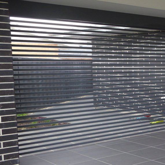 A black garage door with horizontal slats partially closed over a paved area and dark brick wall.