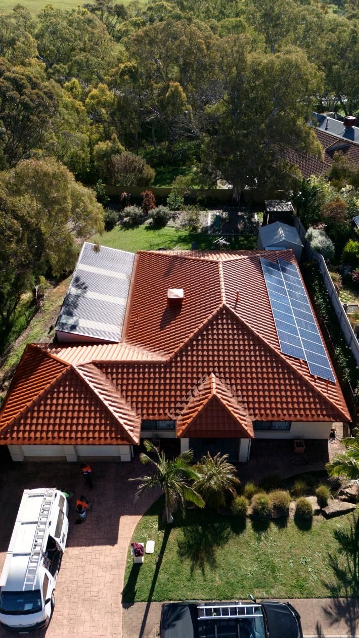 Aerial view of a suburban house with a red tile roof, solar panels, and a driveway beside trees.