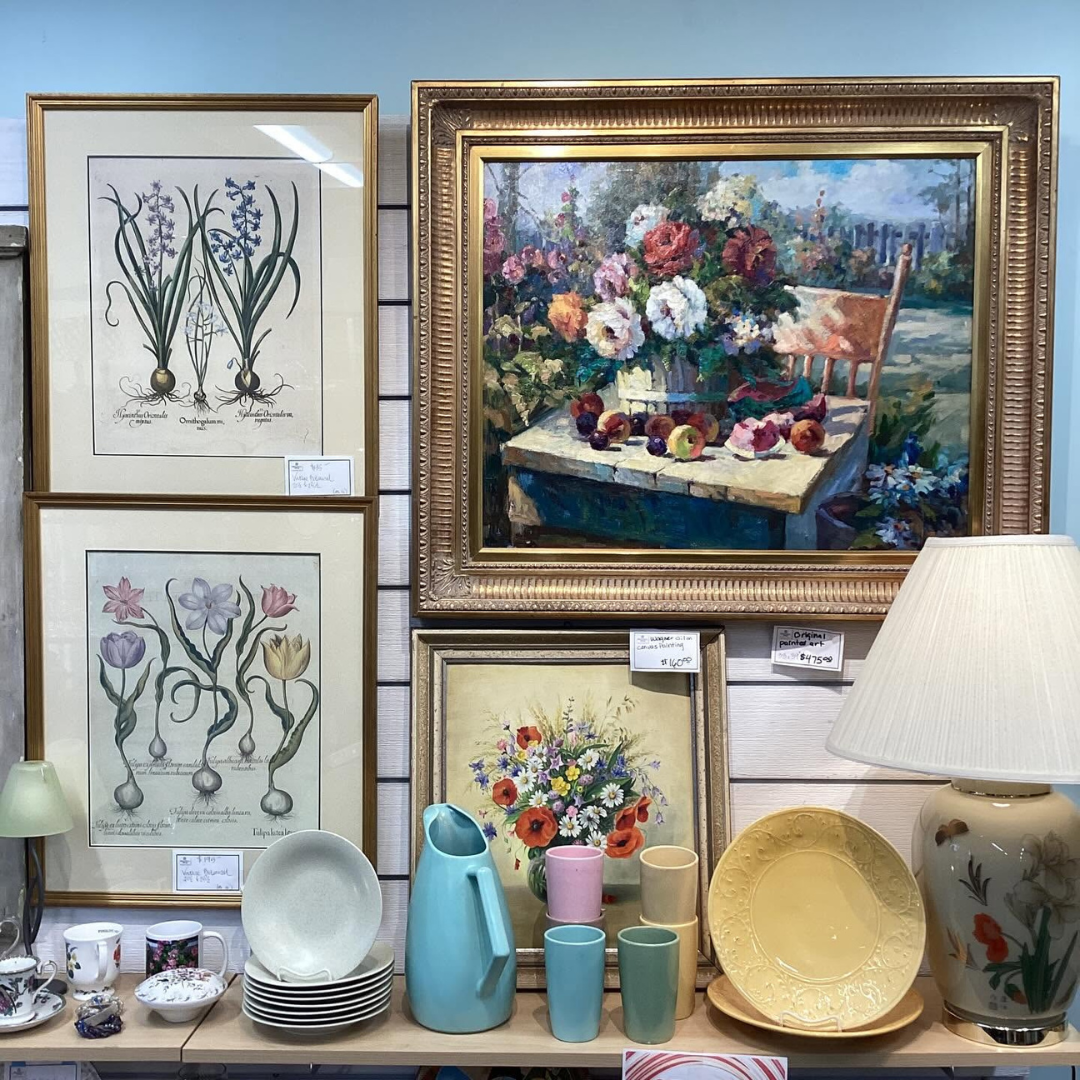 Assorted framed floral paintings, ceramics, and tableware displayed in an art shop window