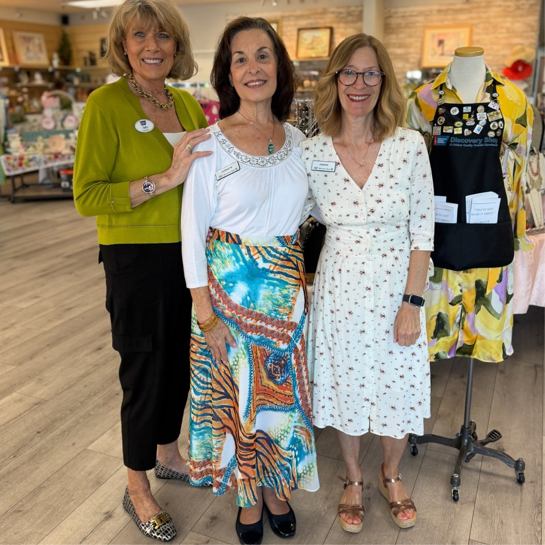 Three women posing in a boutique, wearing colorful dresses and smiling beside a mannequin.