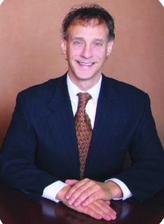 Man in navy suit, brown tie, smiling, seated at a desk with clasped hands.