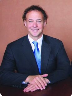 Man in a suit smiles, hands clasped. He wears a blue tie, sitting at a table against a brown backdrop.
