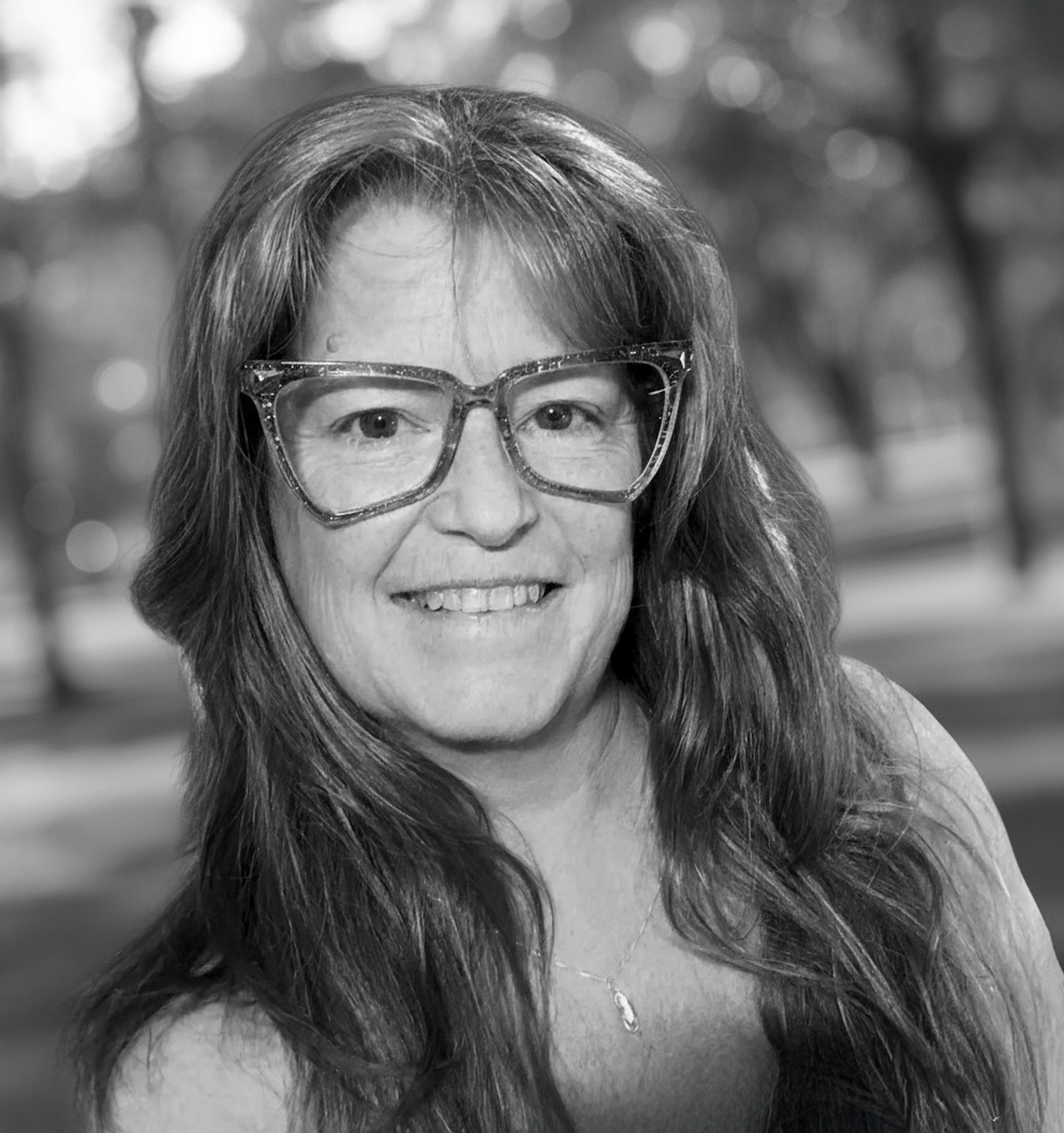 Woman with long hair wearing glasses smiling in an outdoor setting.