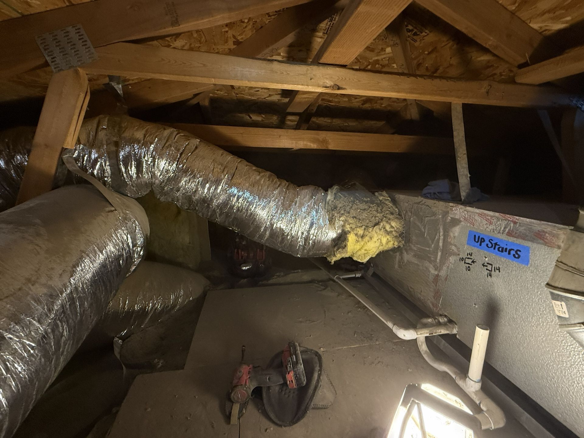 Attic crawlspace with insulated ducts, exposed wooden beams, and a cordless drill on the floor.