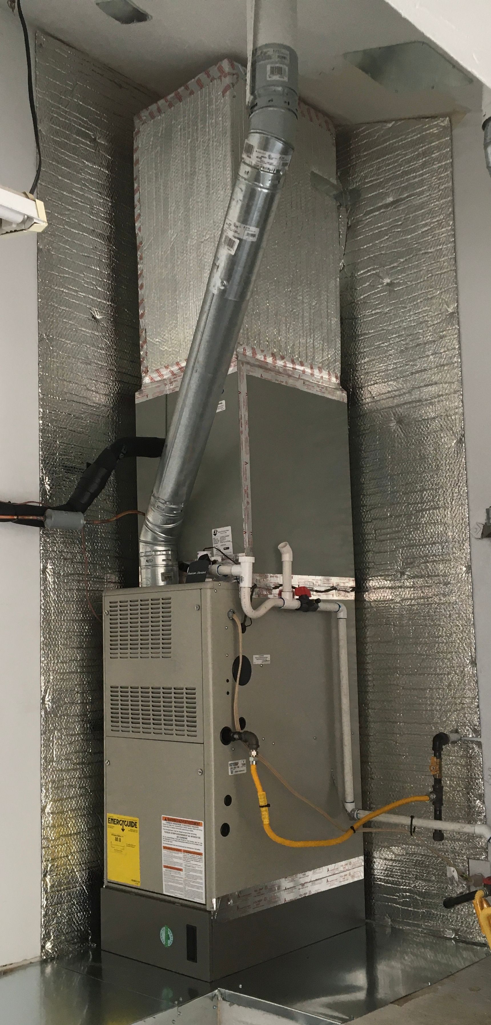Industrial HVAC furnace with silver ductwork in a utility closet, surrounded by insulated walls.
