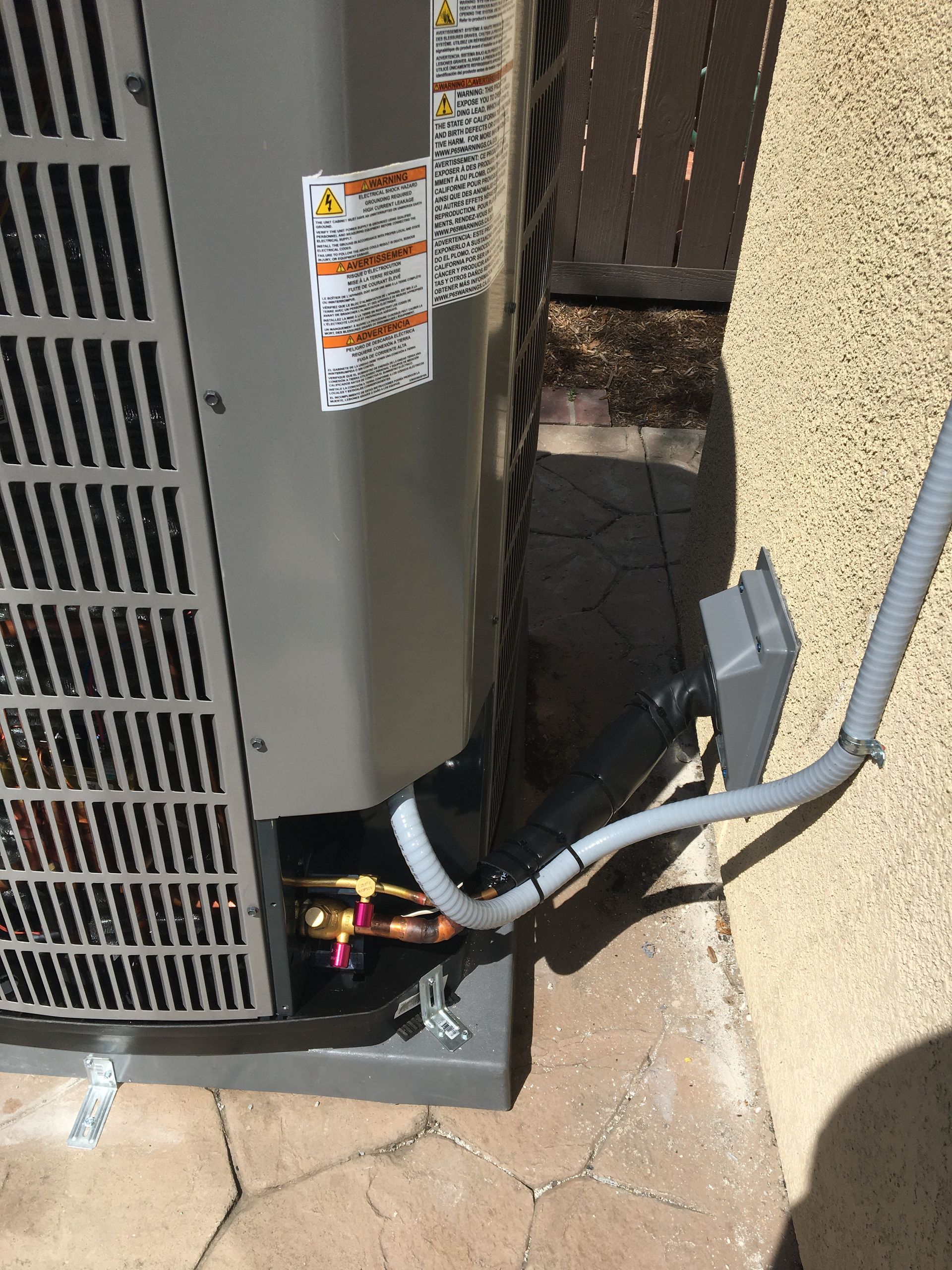 Outdoor HVAC unit with exposed electrical wiring beside a stucco wall and walkway