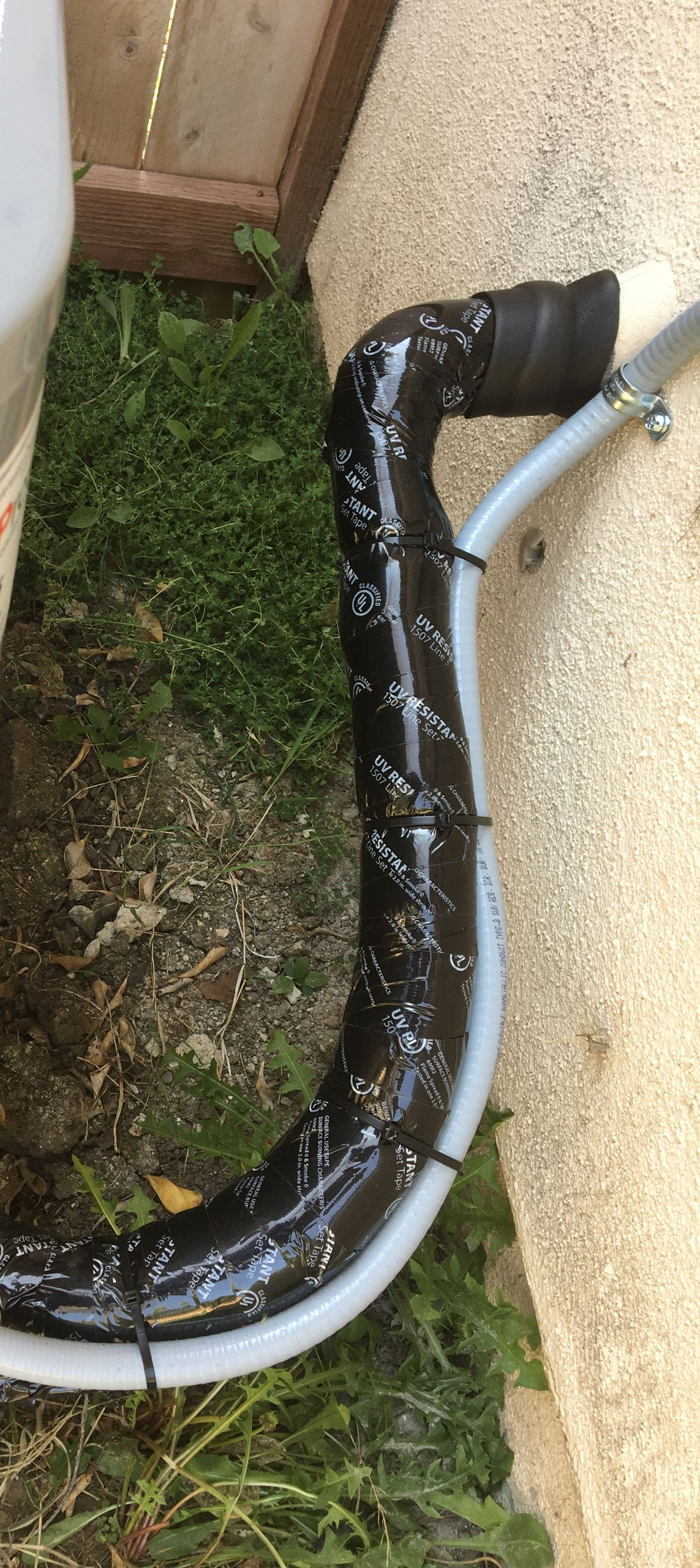Black pipe wrapped in duct tape running along a wall and grass outdoors