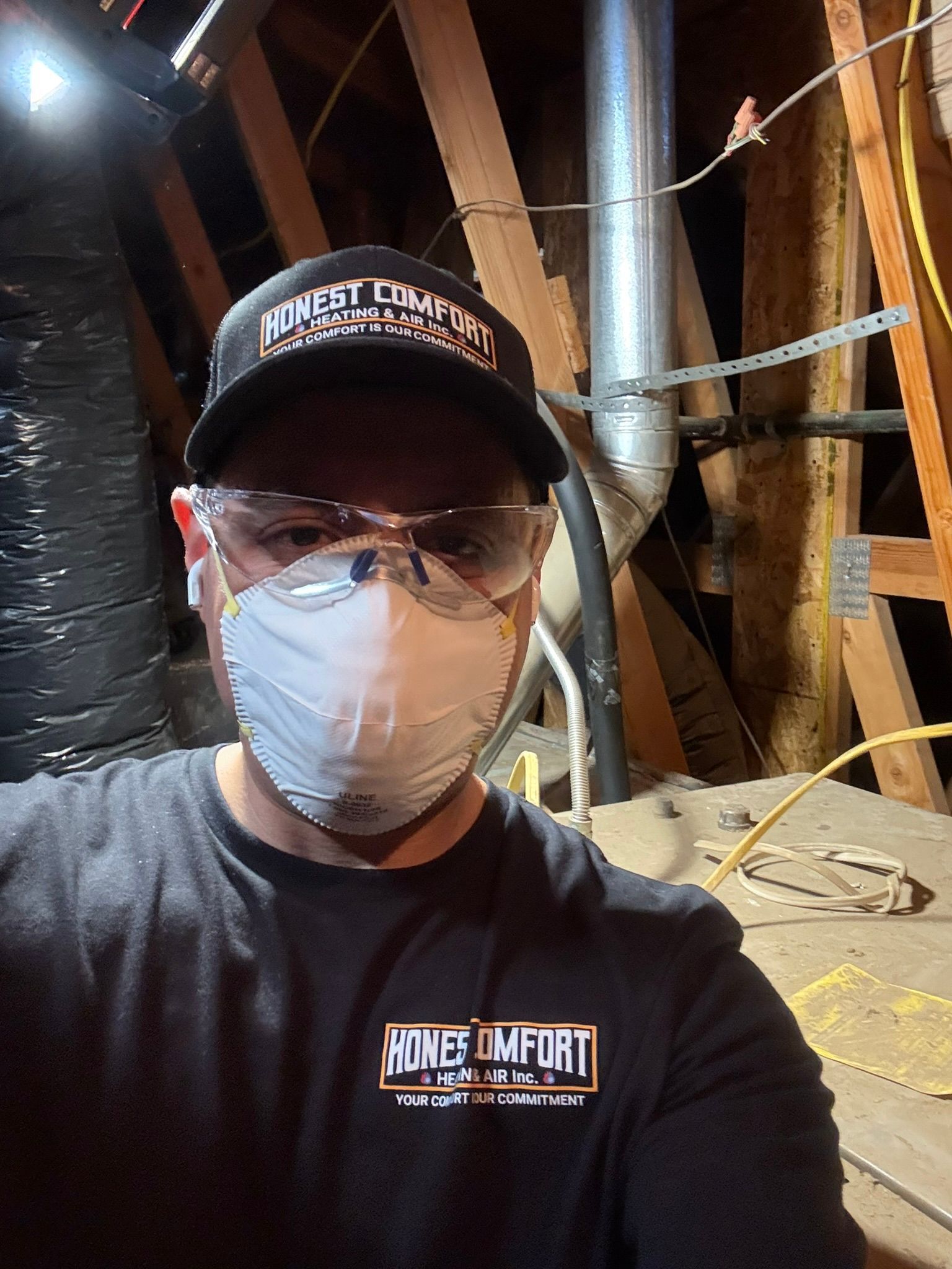 Person in a black cap, glasses, and mask taking a selfie in an unfinished attic with exposed pipes and beams.
