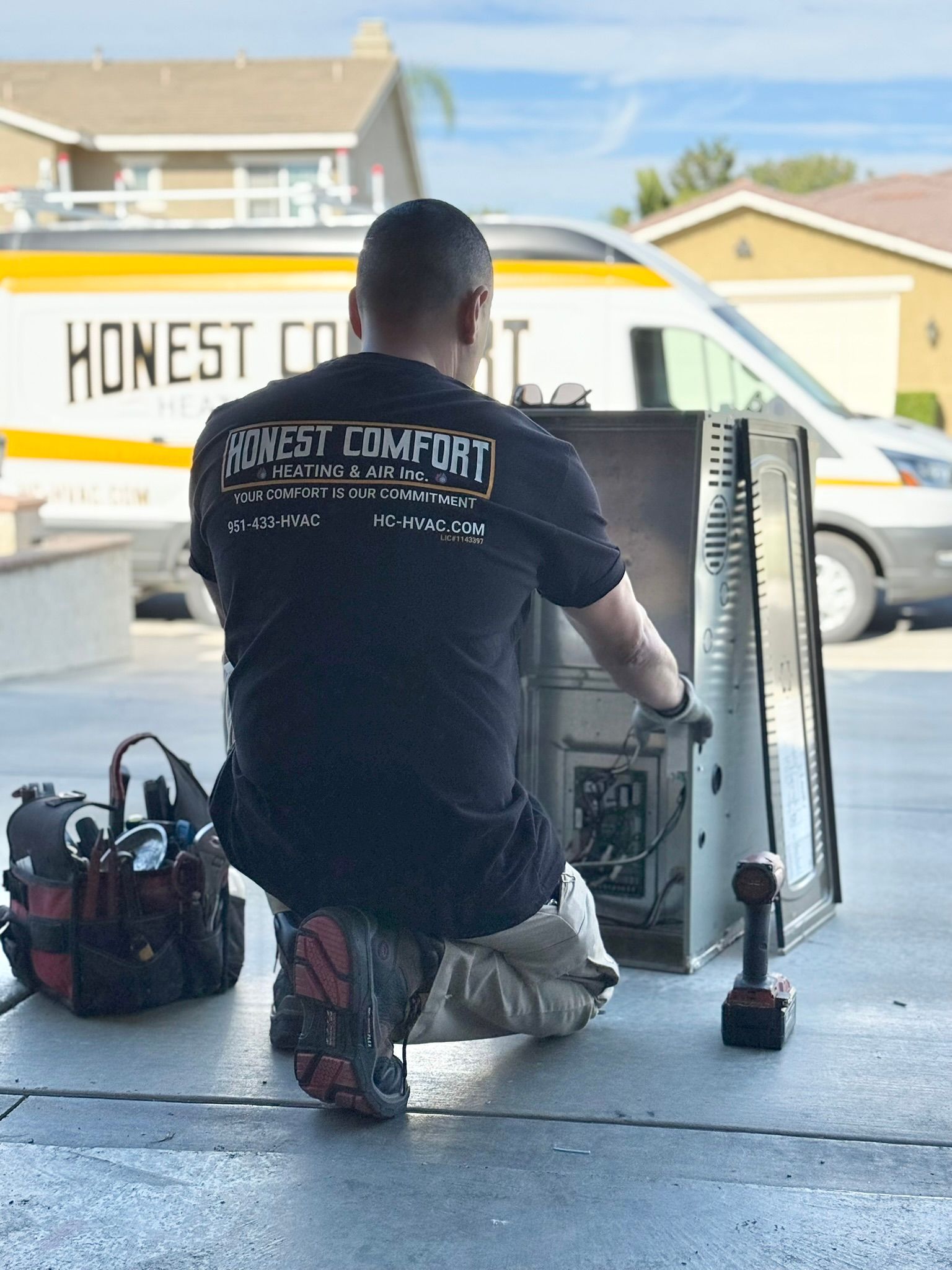 Worker kneels beside open HVAC unit in a garage, using tools on a sunny driveway.