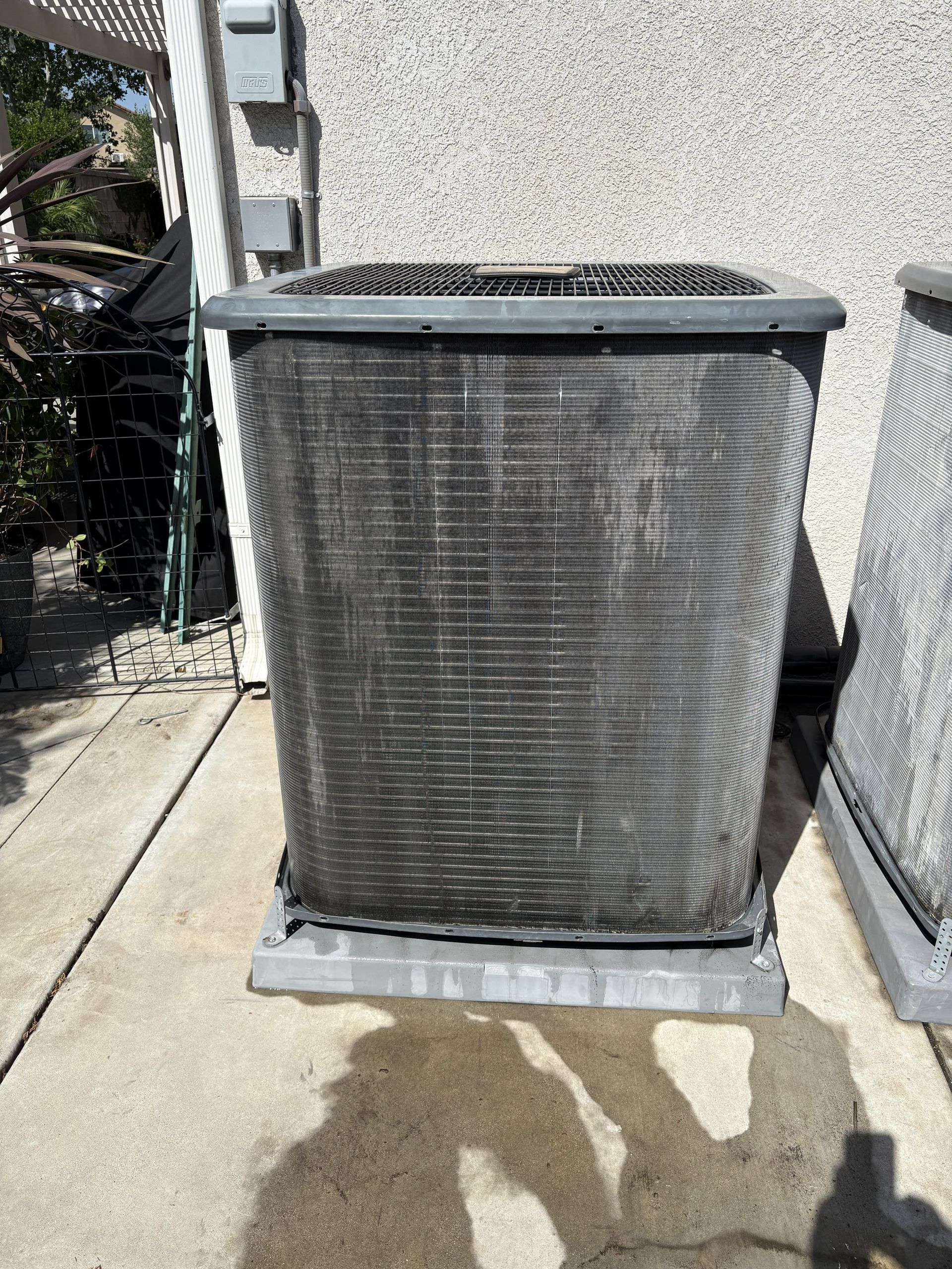 Outdoor HVAC condenser unit against a stucco wall on a concrete pad, with a fence and shadows nearby