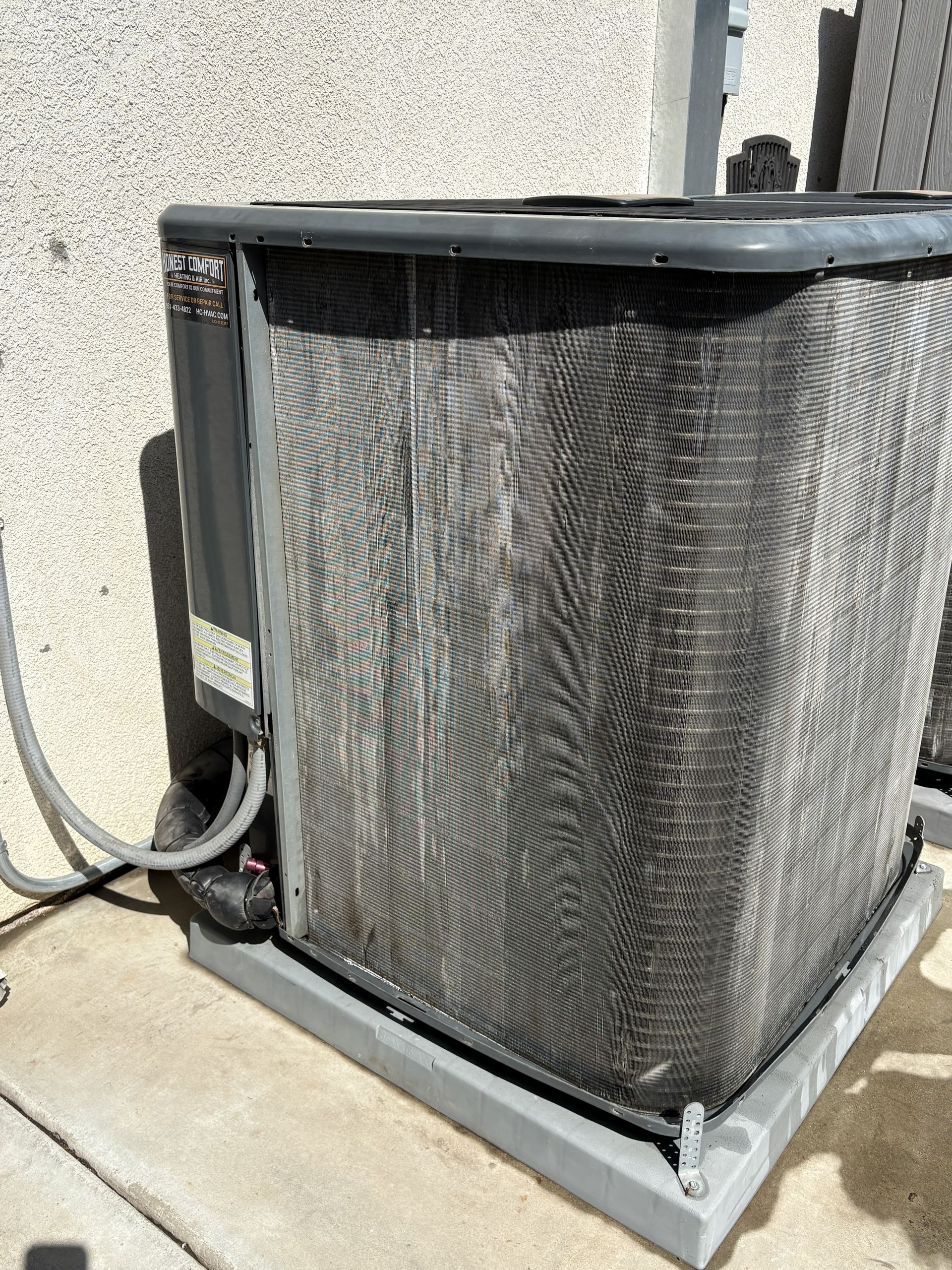 Outdoor HVAC condenser unit with a black top, gray mesh sides, and connected refrigerant lines on a concrete pad